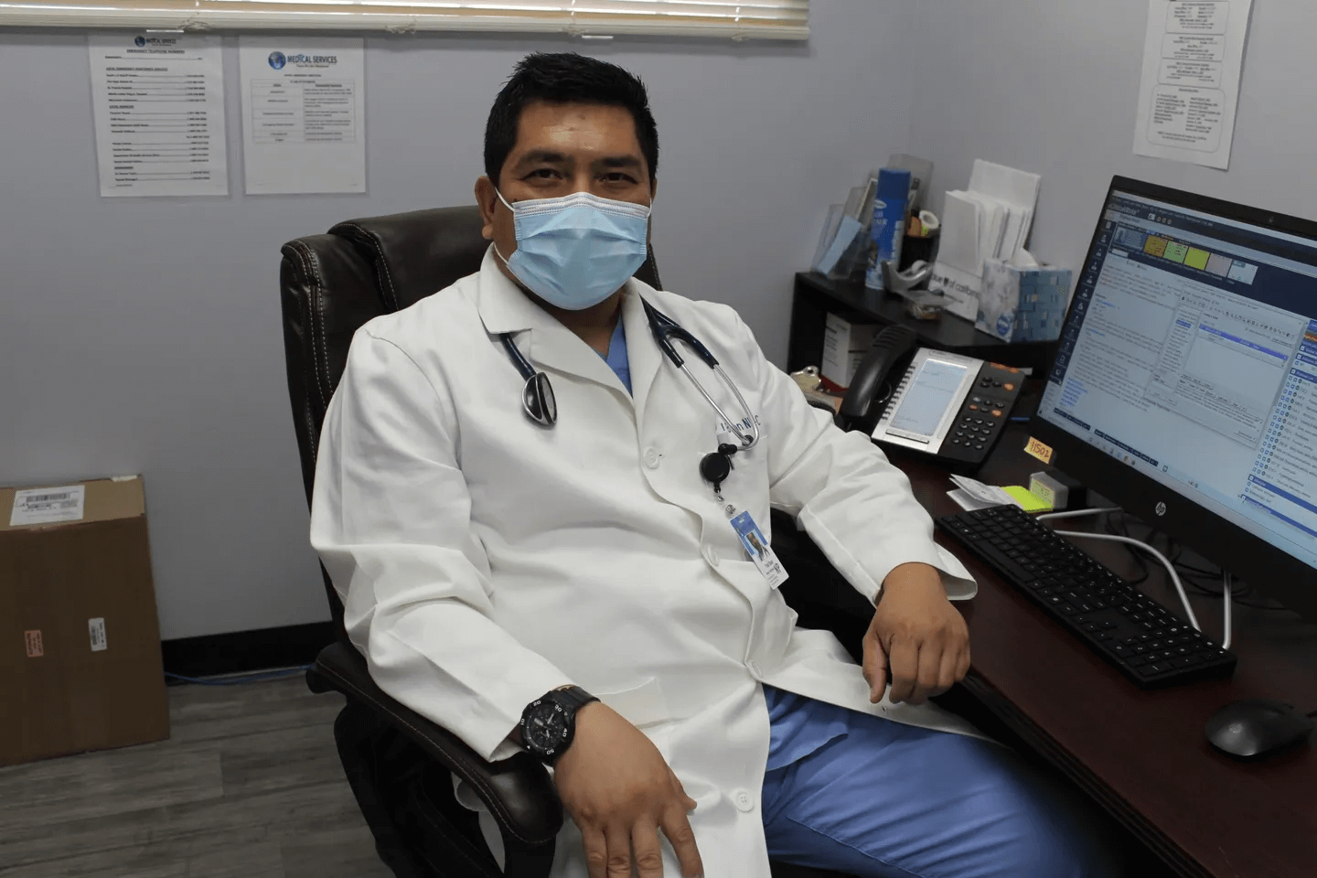 A doctor sitting in his office wearing a mask.